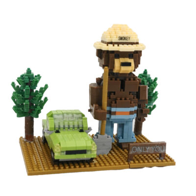 A completed Smokey Bear mini building blocks set featuring Smokey Bear standing on a forest-themed base with trees and a green car beside him. Smokey is holding a shovel, wearing his iconic ranger hat labeled 'Smokey,' and standing next to a sign that reads 'Only You.' The assembled model consists of small, colorful building blocks with a detailed design.