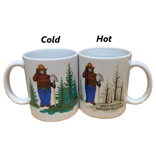 Two Smokey Bear Magic Mugs side by side, showing the color change from a green forest when cold to a post-wildfire landscape when hot, with Smokey Bear and the message 'Only you can prevent wildfires' visible on both.
