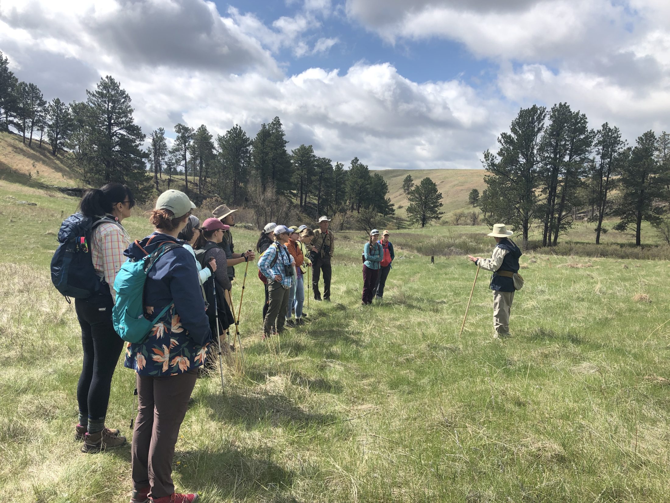 A group of people on a guided hike. It is spring, there are ponderosa pine trees and green grass, with large puffy clouds in the sky. The instructor is a female wearing a tan hat, with a navy blue jacket