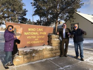 2 men and 1 women standd near the Wind Cave National Park sign. It is winter they are happy to be there and are wearing winter coats.