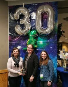 Three women stand in front of a banner that has a thirty and dinosaur ballon. They are dressed in buisness causal at an event hall. They are holding an award that says "Friend of Science"