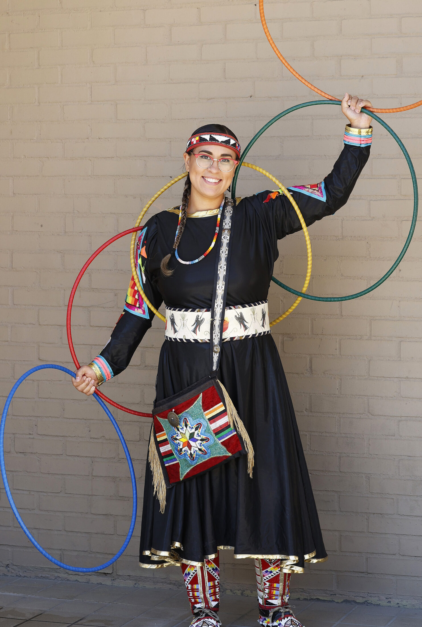 Woman wearing a black dress with beadwork on her dress and moccasins. She is holding four colorful hoops while standing in front of a tan brick wall.