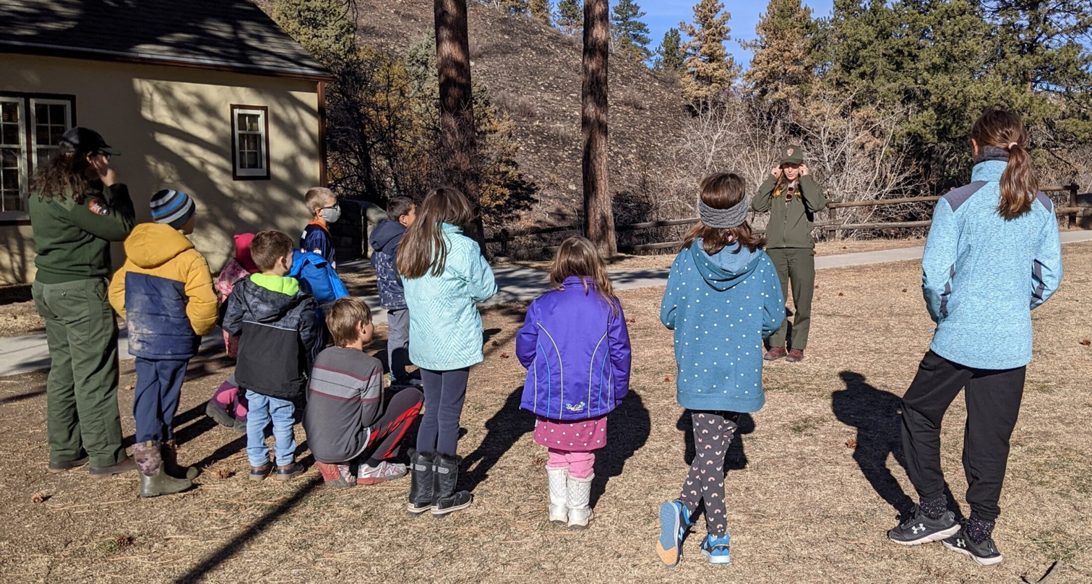 Young children with their backs to the camera dressed in winter coats standing on a brown lawn looking at a ranger in a green uniform who is talking to them. In the background is the corner of a yellow building with trees and a black hillside in the center of the photo.