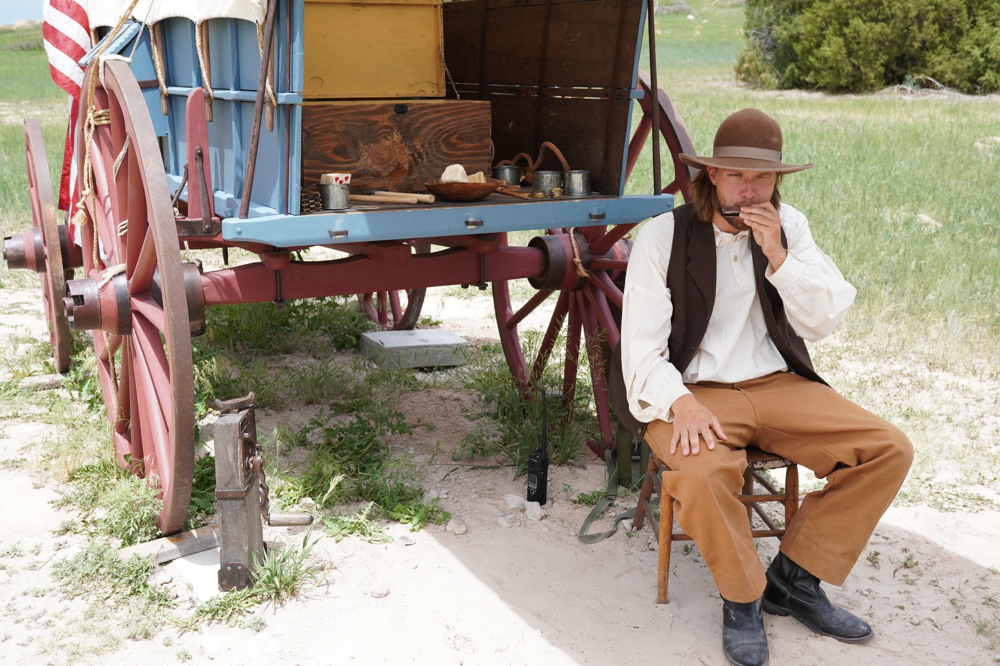 White male in white linen shirt, dark vest and brown pants appearing to be from the 1850s sits behind a blue painted covered wagon.  He plays the harmonica.