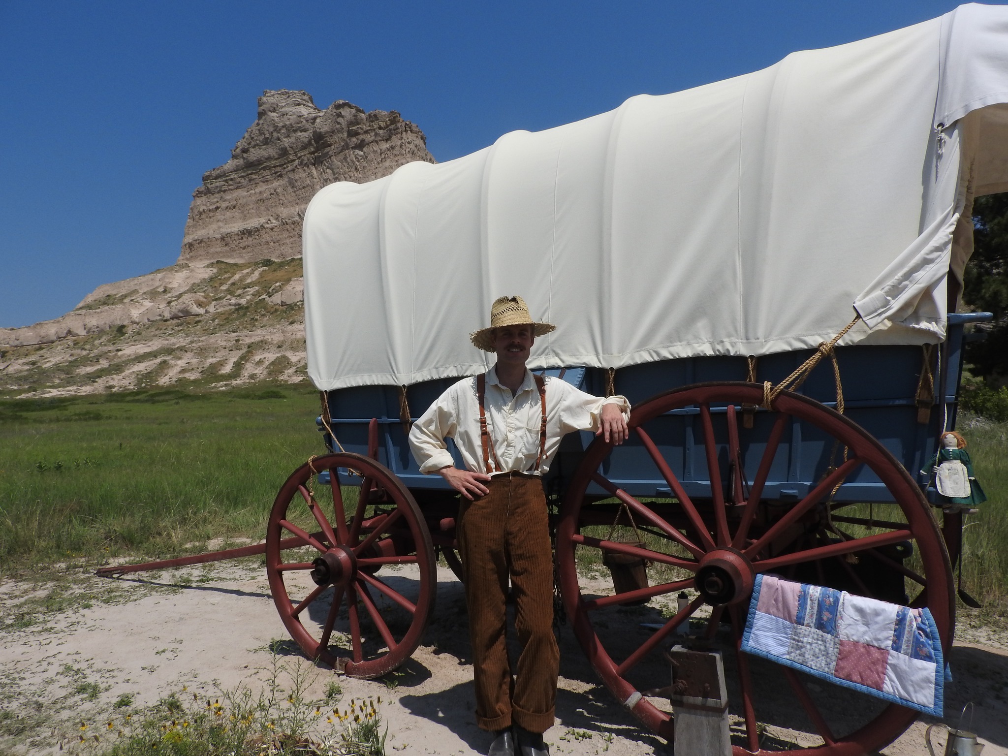 Tall thin white male in linen 1850s style clothing and a hat stands in front of a brown covered wagon with large wood wheels on a pale gray gravel surface. A bright blue sky and a tall bluff hillside in the background.