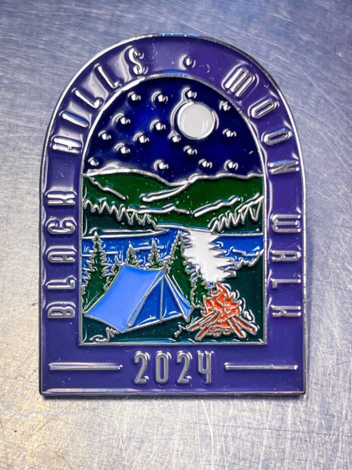 Colorful metal lapel pin lying on a silvery metal tabletop. Text on pin in arch shape reads "Black Hills Moon Walk - 2024". Center of pin is a night scene with a blue tent and orange campfire in the foreground, blue lake reflecting a silver moon in the background.