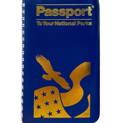 Close-up image of the Passport To Your National Parks® Classic Edition cover. The cover is a deep blue with gold embossed text and graphics, featuring an eagle in flight and a waving American flag with stars. The spiral binding is visible along the left side.