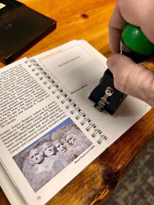 Close-up of a hand holding a green and black rubber stamp, stamping a cancellation mark onto a page in a spiral-bound Passport To Your National Parks® book. The book is open to the Rocky Mountain Region section and features a photo of Mount Rushmore and text describing its history. The scene is set on a wooden table.