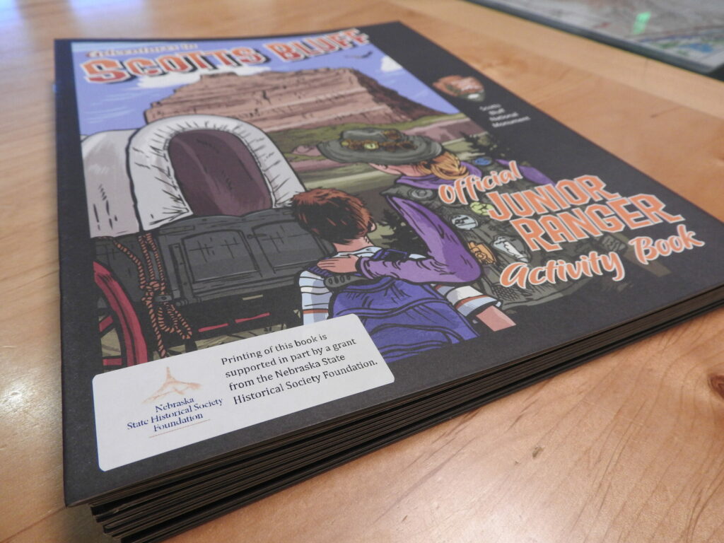 Thin booklet with a cartoon covered wagon lies on a wooden desk. A label with the NSHSF logo is prominent in the lower left corner of the front page