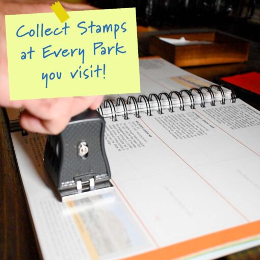 Close-up image of a person stamping a page in a spiral-bound national park passport book. The page has sections for different parks, each with descriptions and space for stamps. A yellow sticky note graphic is overlaid on the image, with text that reads, "Collect Stamps at Every Park you visit!" The scene captures the excitement of documenting visits to various national parks by collecting unique cancellation stamps.