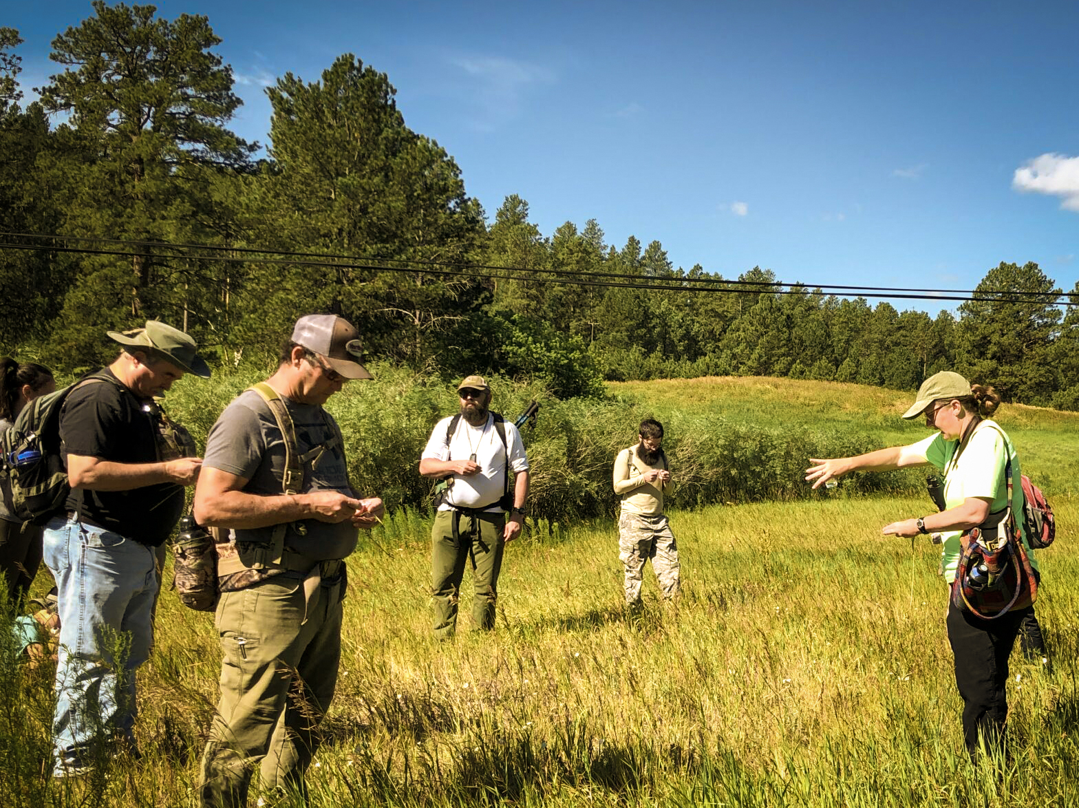 A group of people in outdoor trail maintenance gear work in a grassy field under a blue sky with puffy white clouds.