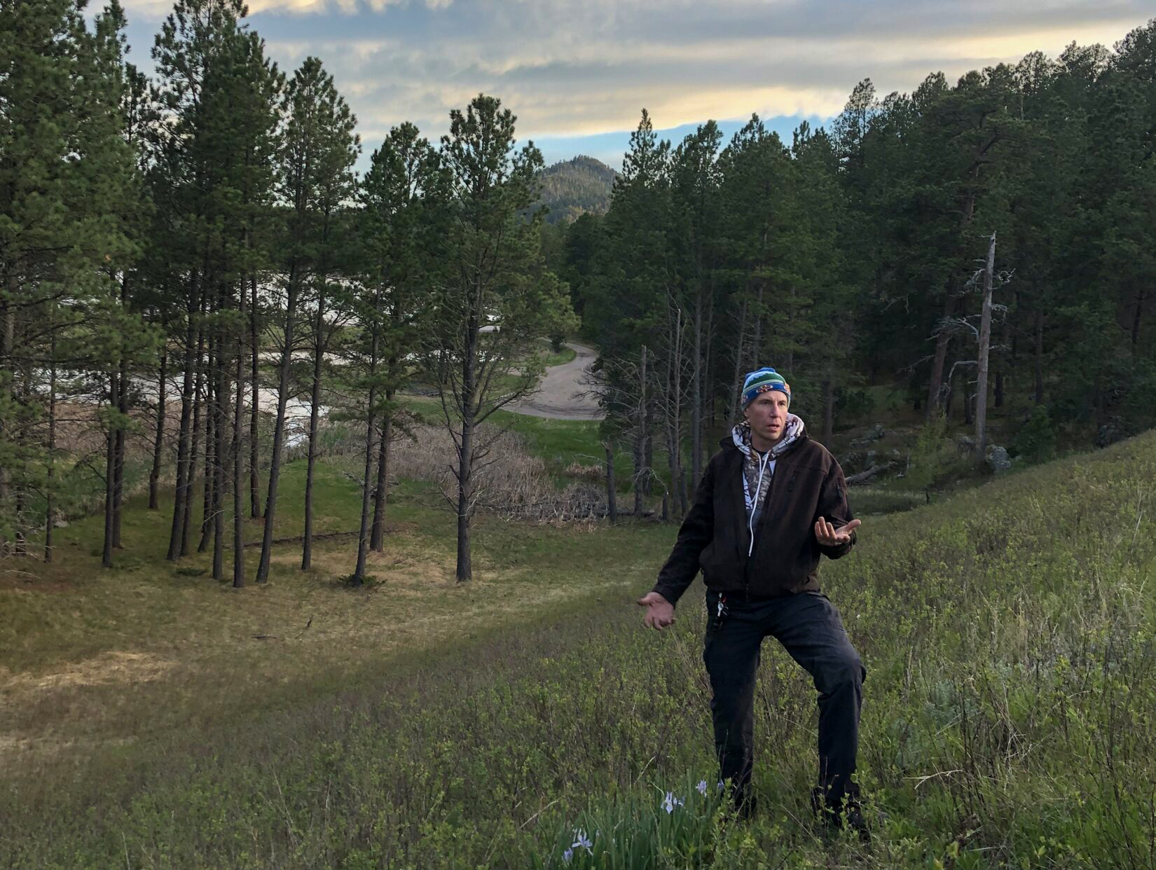 Man wearing dark jacket and pants gestures towards camera in front of a forest valley with dramatic sunset beyond.