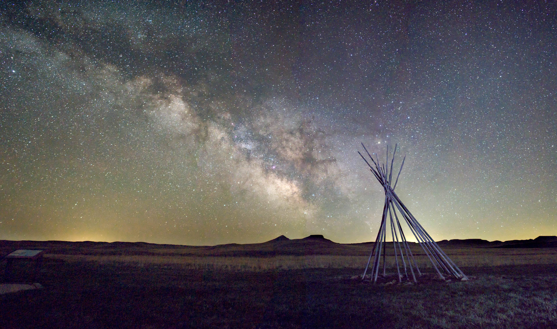 Starry night sky with the milky way extending from lower right to upper left. Tipi poles in the shape of a lakota lodge in the near foreground. 