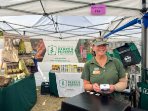 photos of the interior of white fabric-topped canopy tents. Two customers hold an open book while speaking with a man in green shirt and dark ballcap. Woman in green logo shirt with green ballcap holds a credit card machine while smiling at the camera. Merchandise and BHPFA logo signage in background.