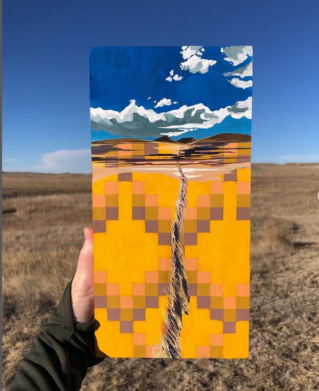 A hand holds up a rectangular painting against the backdrop of an open, grassy landscape under a clear blue sky. The painting features a stylized depiction of the same landscape, with geometric patterns in vibrant yellow, orange, and pink tones representing the ground, and a deep blue sky with white clouds above. A path or crack runs through the center of the image, creating a sense of depth and movement. The painting draws inspiration from the natural surroundings, blending traditional and modern artistic styles.