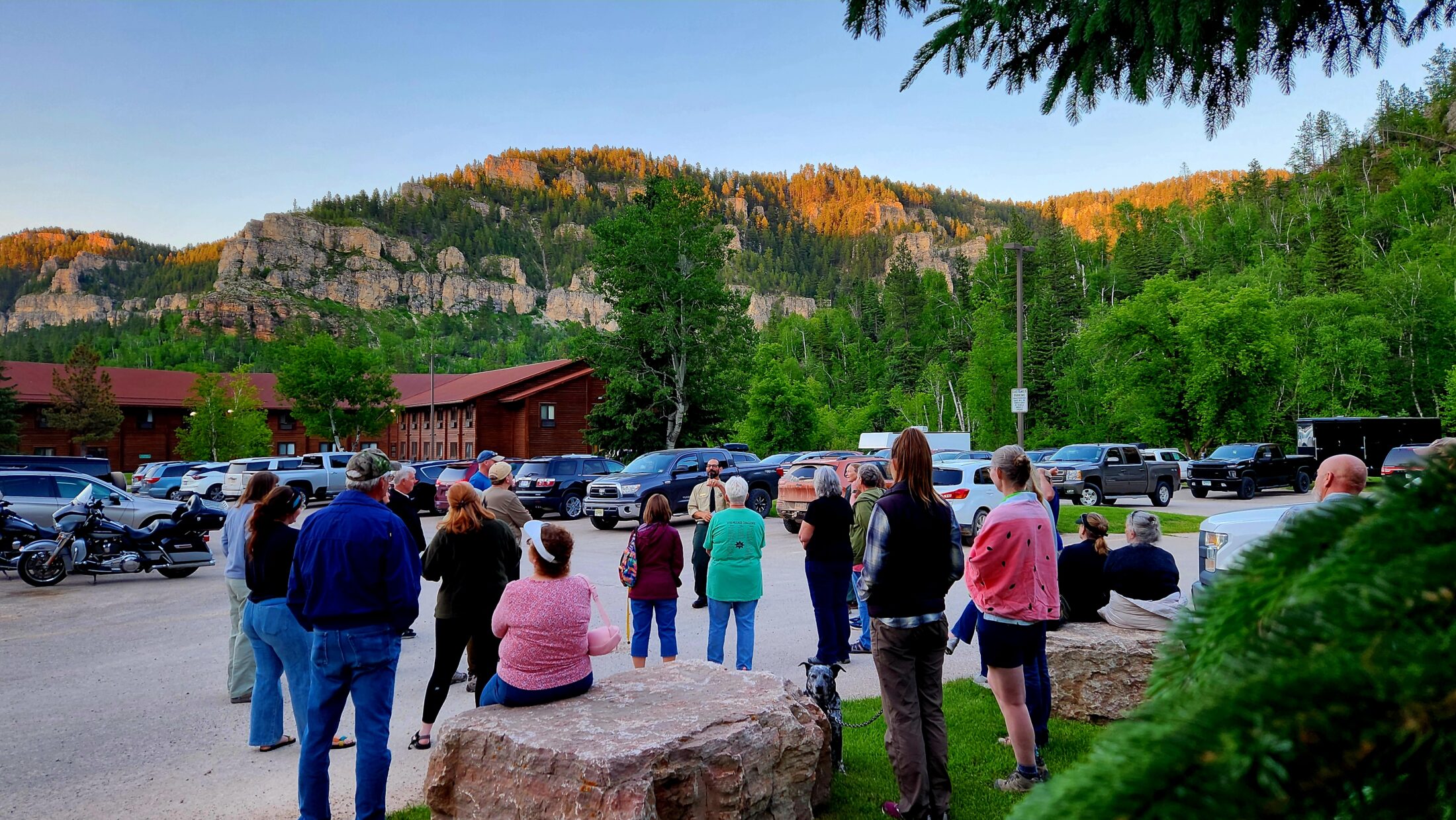 A group of people standing and sitting on large rocks, gathered outdoors in a parking lot surrounded by cars and motorcycles. They are listening to a park ranger, who is speaking in front of them while wearing a uniform and hat. The group is in a scenic area with tall cliffs and a dense forest in the background, bathed in the warm glow of a setting sun. In the distance, a large, rustic lodge is nestled among the trees at the base of the cliffs. The surrounding landscape is filled with lush greenery, and the cliffs are partially illuminated by the fading sunlight