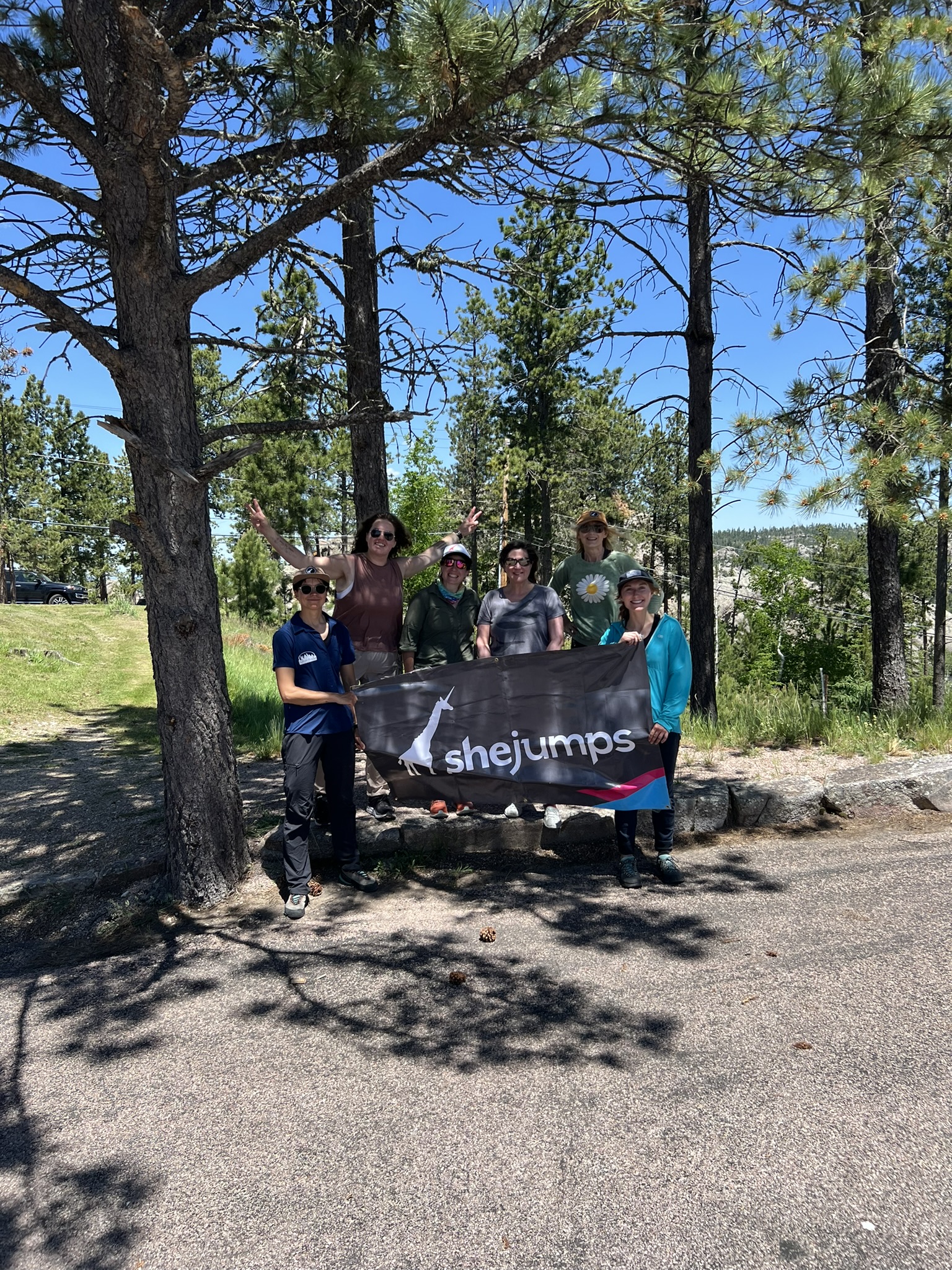 A group of six women stands outdoors in a forested area, holding a banner that reads "SheJumps" with the organization's logo of a white giraffe. The women are smiling and dressed in casual outdoor clothing, with some wearing hats and sunglasses. They are surrounded by tall pine trees, and the bright blue sky overhead indicates a sunny day. The group stands on a paved area, with dappled sunlight casting shadows on the ground. The scene reflects a cheerful and empowering outdoor gathering.