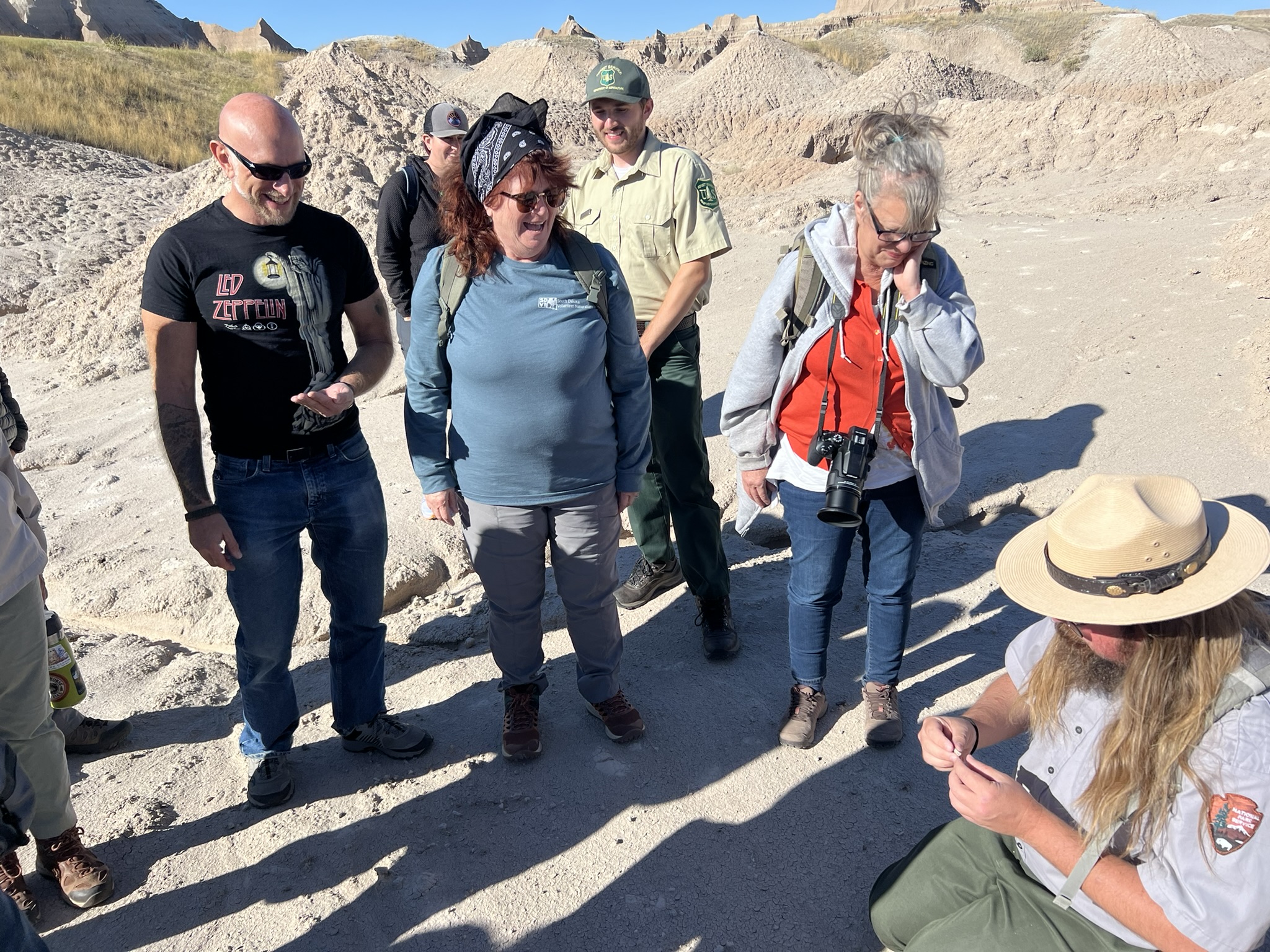 A group of people stands in a rocky, desert-like area, watching a National Park Service ranger who is kneeling and holding a small fossil. The ranger, with long hair and a wide-brimmed hat, is showing the fossil to the group. The onlookers are dressed for outdoor exploration, with some wearing hats, sunglasses, and backpacks. One person has a camera hanging around their neck. The background features rugged, eroded hills and rock formations under a clear blue sky, typical of a Badlands landscape.