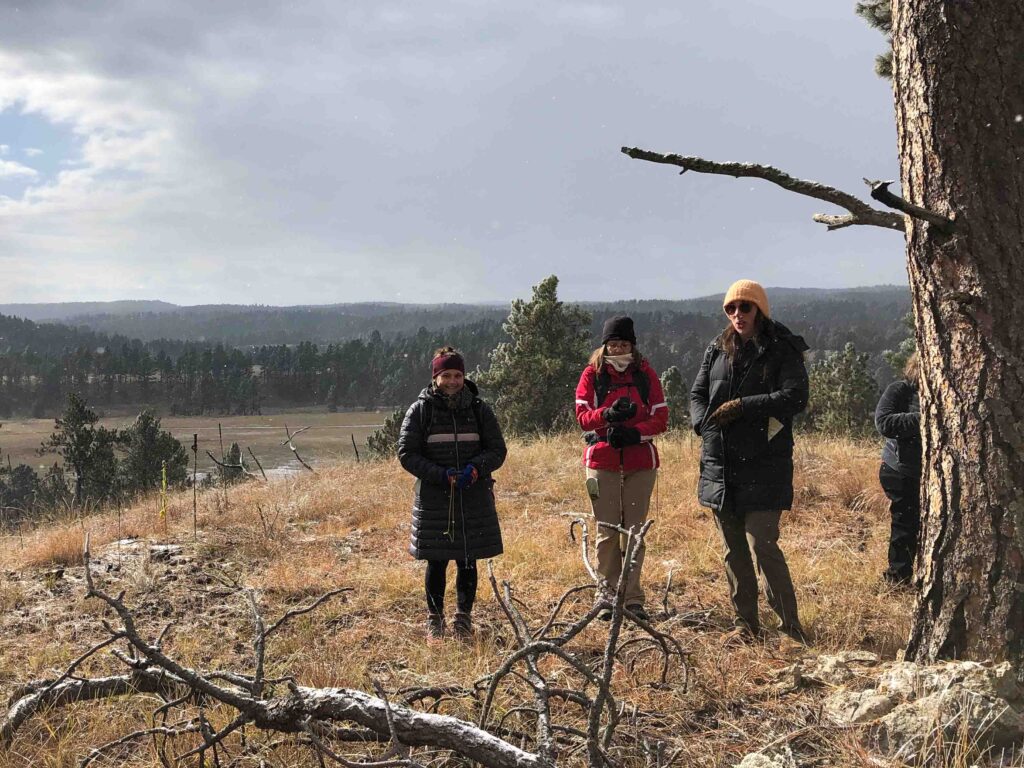 Three women dressed in winter outdoor clothing stand on a grassy hilltop under a cloudy sky. They are practicing map and compass navigation skills. A large tree trunk is visible on the right, and a scenic view of rolling hills and distant forests stretches out behind them.