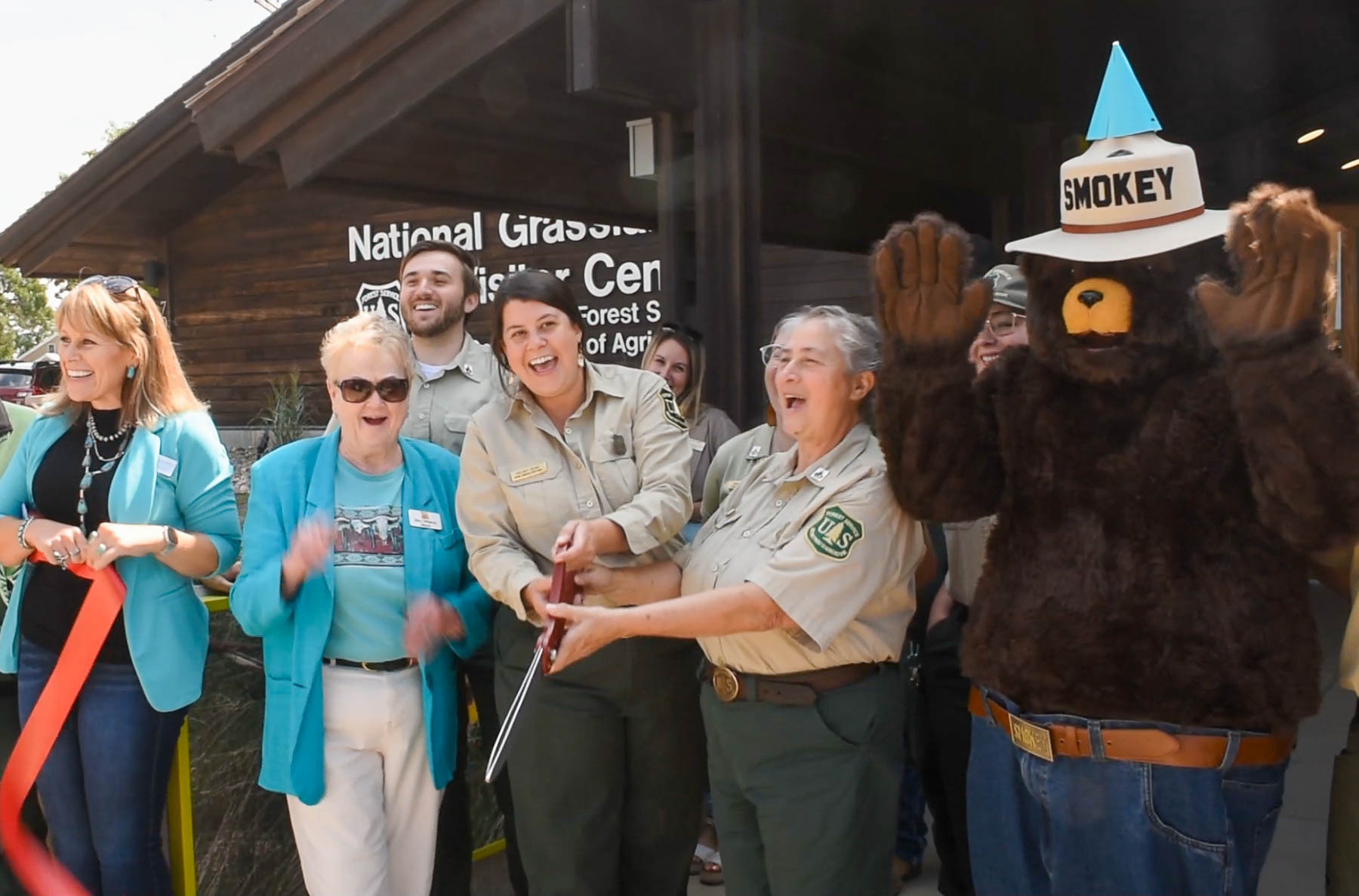 A group of people celebrates a ribbon-cutting ceremony in front of a building marked "National Grasslands Visitor Center." A woman in a Forest Service uniform cuts the ribbon with large scissors, while others around her smile and cheer. Smokey Bear, wearing his signature hat and a blue party hat, stands to the right with his arms raised. The group includes people in Forest Service uniforms and others in casual or business attire, creating a lively, celebratory atmosphere on a sunny day.