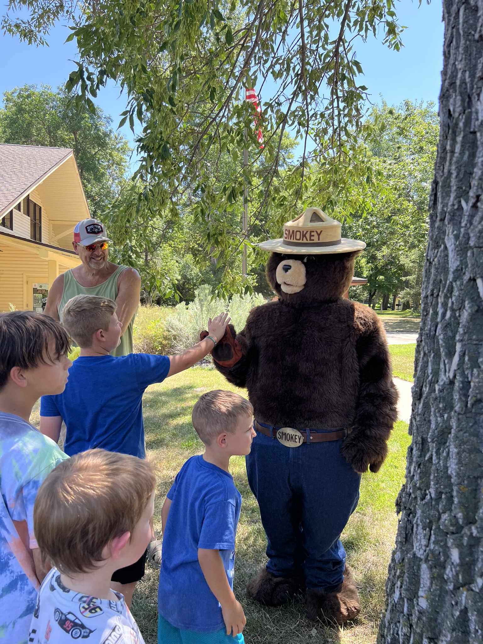 Smokey Bear, wearing his signature ranger hat and blue jeans with a belt buckle that says 'Smokey,' gives a high five to a young boy in a group of children. The children are gathered around Smokey on a grassy area under the shade of a large tree. In the background, a man in a tank top and sunglasses watches the interaction, smiling. An American flag is visible in the distance, along with a yellow house nestled among trees. The scene takes place on a sunny day, with lots of greenery surrounding the area.