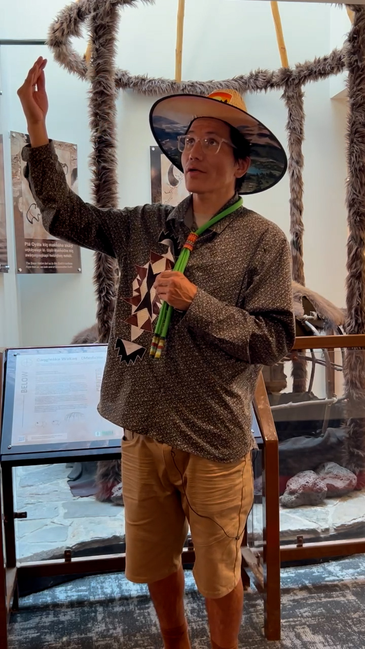 A man wearing a wide-brimmed hat, a patterned shirt, and shorts speaks to an audience inside a museum exhibit. He holds a microphone in one hand and gestures upward with the other. The exhibit behind him features large animal pelts and artifacts, with informational plaques and displays. The man appears to be engaging the audience in a storytelling or educational presentation, with a calm and focused expression. The setting is indoors, with soft lighting highlighting the historical items on display.<br />
