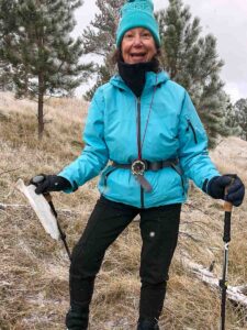 A woman smiles brightly while holding a compass in one hand and a map in the other. She is dressed in a bright blue winter jacket, black pants, and a turquoise beanie, standing among trees and snow-dusted grass. She appears to be enjoying her map and compass navigation practice.