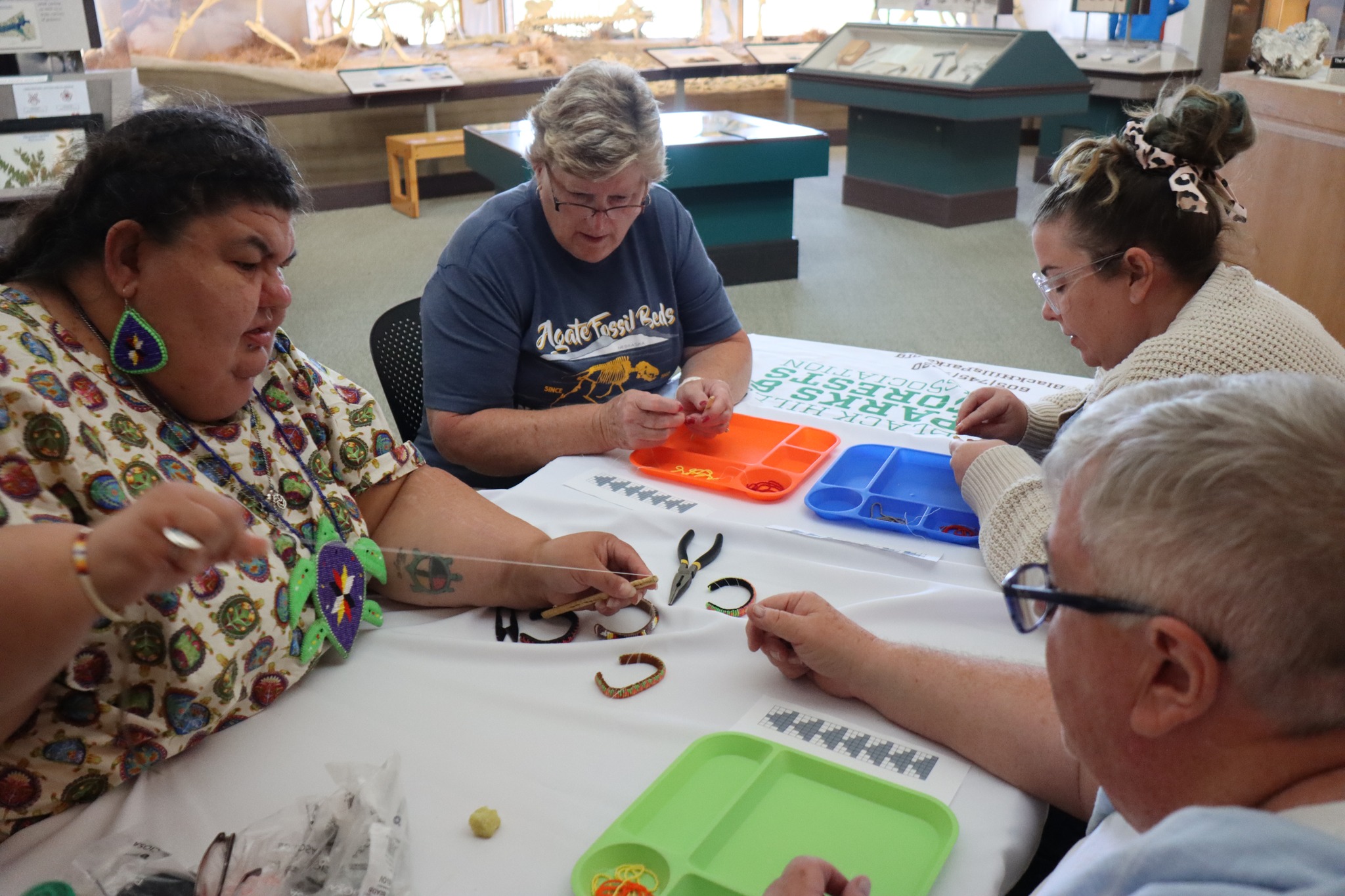 Four people sitting around a table inside a museum space, engaged in crafting activities. On the left, a woman wearing a colorful, patterned top and large beaded turtle jewelry works on a project with pliers and beading materials. In the center, a woman wearing an 'Agate Fossil Beds' T-shirt focuses on her craft, and on the right, a younger woman with glasses and a hair scrunchie leans over her work. A man with short white hair and glasses is also participating, working on the project in front of him. Various tools, beads, and materials are spread out on the table, along with colorful trays and a large banner featuring the Black Hills Parks and Forests Association logo. Museum exhibits, including fossils and displays, are visible in the background