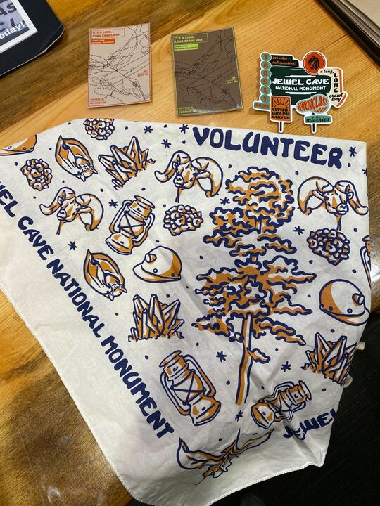 A display of volunteer appreciation items from Jewel Cave National Monument. In the center is a white bandana with blue and orange illustrations, including a tree, lanterns, crystals, bats, and helmets, along with the words "Jewel Cave National Monument" and "Volunteer." Above the bandana are two topographic map postcards and a set of colorful Jewel Cave-themed stickers. The items are laid out on a wooden surface.