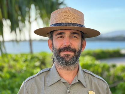 A park ranger stands outdoors, wearing a traditional straw ranger hat and a beige National Park Service uniform. He has a full beard, and the background features lush green foliage and a body of water under a clear blue sky, creating a tropical or coastal setting. His expression is calm and welcoming, framed by the natural beauty of the surroundings.