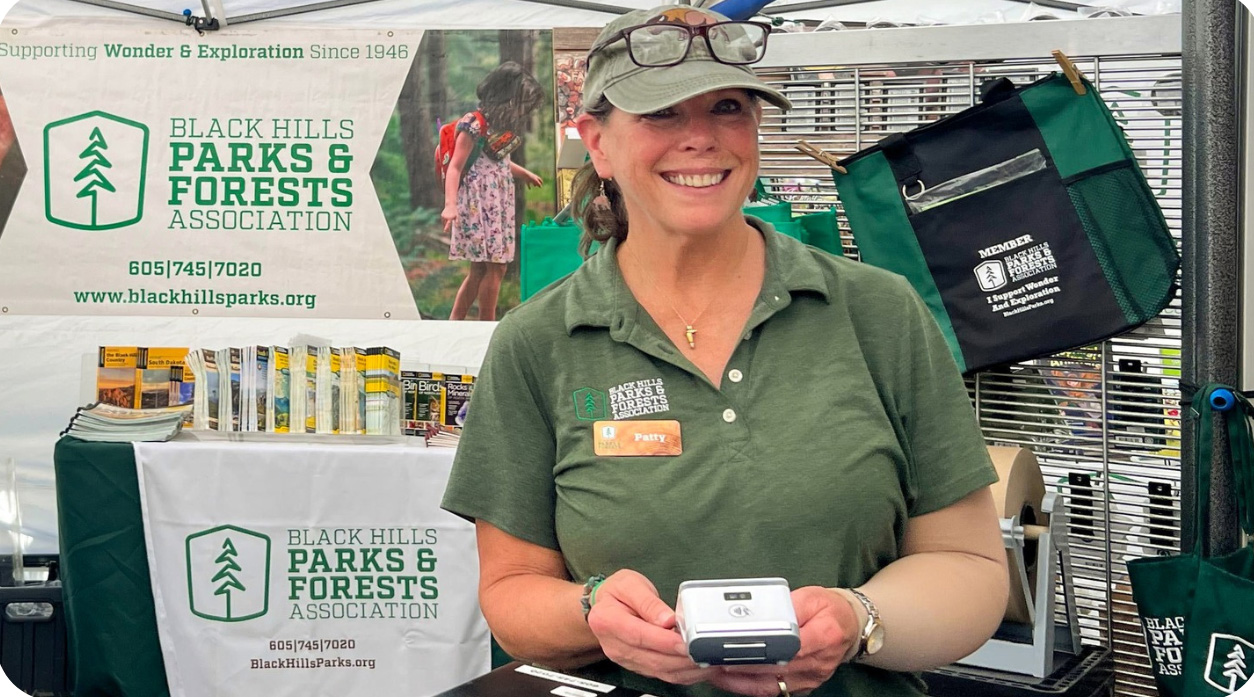 A woman wearing a green polo shirt with a "Black Hills Parks & Forests Association" logo and a name tag reading "Patty" stands at an informational booth. She is smiling and holding a small device in her hands. Behind her, the booth displays a banner with the association's logo and contact information, as well as promotional materials, including brochures and tote bags. The setting appears to be an outdoor event or market, with the focus on engaging with visitors about the association.