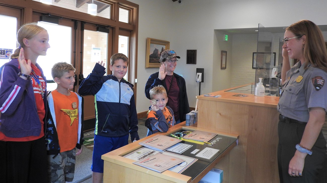 A National Park Service ranger administers the Junior Ranger oath to a group of children and an adult in a park visitor center. The children and adult hold up their right hands while one child shows off a Junior Ranger badge. The ranger stands behind a wooden desk with certificates and Junior Ranger booklets on display. Sunlight shines through large windows in the background. Everyone is smiling, capturing a moment of fun and learning at the park.