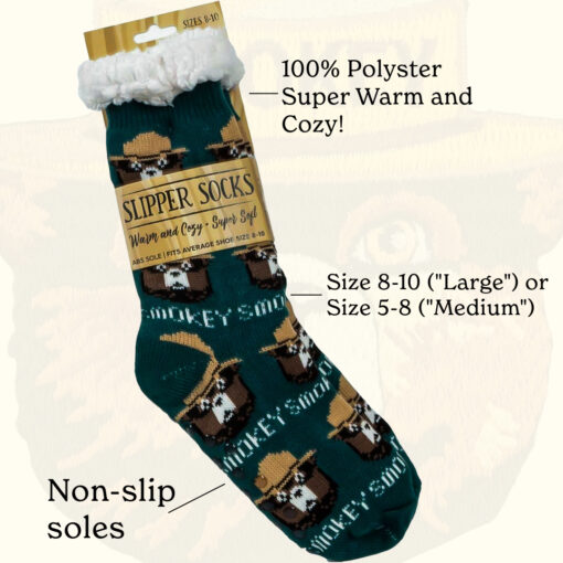 A pair of green Smokey Bear-themed slipper socks with detailed feature labels pointing to '100% polyester,' 'super warm and cozy,' 'non-slip soles,' and sizing options ('Large' for size 8-10 and 'Medium' for size 5-8). The socks have a fluffy white cuff and a repeating Smokey Bear pattern wearing his iconic ranger hat."