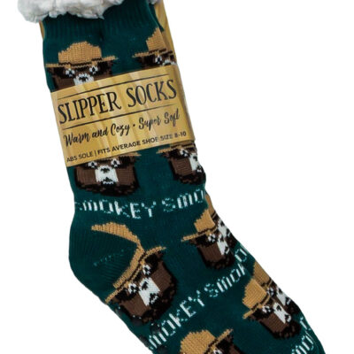 Close-up of Smokey Bear-themed slipper socks packaged with a tag reading 'Slipper Socks: Warm and Cozy, Super Soft.' The socks are green with a repeating pattern of Smokey Bear in a ranger hat and include a fluffy white cuff at the top.