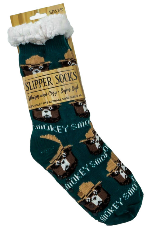 Close-up of Smokey Bear-themed slipper socks packaged with a tag reading 'Slipper Socks: Warm and Cozy, Super Soft.' The socks are green with a repeating pattern of Smokey Bear in a ranger hat and include a fluffy white cuff at the top.