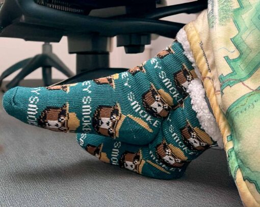 Side view of a person wearing Smokey Bear-themed slipper socks, showing the green fabric with Smokey Bear's face and hat, as well as the soft white cuff. The person's feet rest on a chair leg with a map-patterned blanket partially covering them.