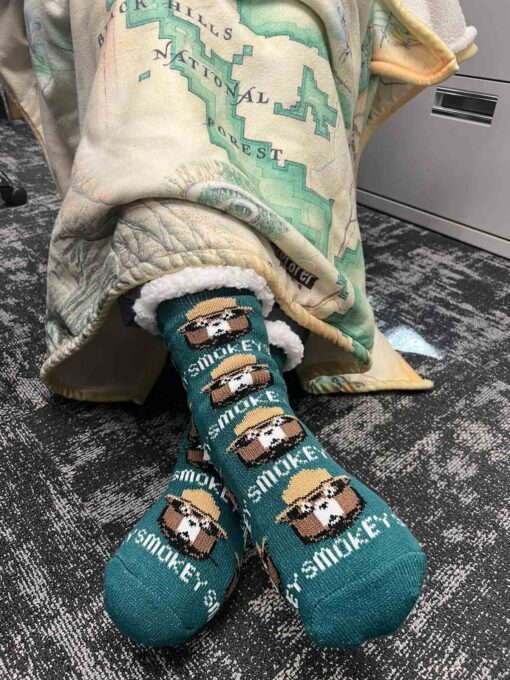 A person wearing Smokey Bear-themed slipper socks with feet stretched forward under a blanket featuring a map of the Black Hills National Forest. The socks are green with Smokey Bear's face in a ranger hat printed across them.