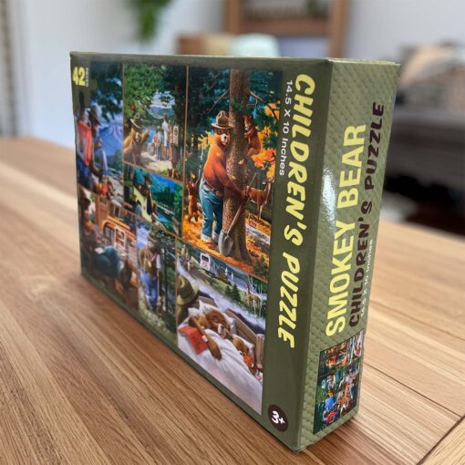 A side view of the Smokey Bear Children's Puzzle box displayed on a wooden table. The box features vibrant artwork of Smokey Bear engaging in forest-related activities, such as protecting wildlife and enjoying the outdoors. The text emphasizes the puzzle's dimensions, 14.5 x 10 inches, and its suitability for children ages 3 and up