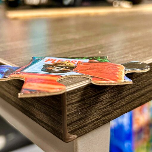 A close-up side view of a single puzzle piece from the Smokey Bear Children's Puzzle. The piece shows part of an illustration with Smokey Bear holding a sign that says 'Please' and includes vibrant red, blue, and green colors. The image also highlights the thick and sturdy cardboard material of the puzzle piece, resting on the edge of a wooden table.