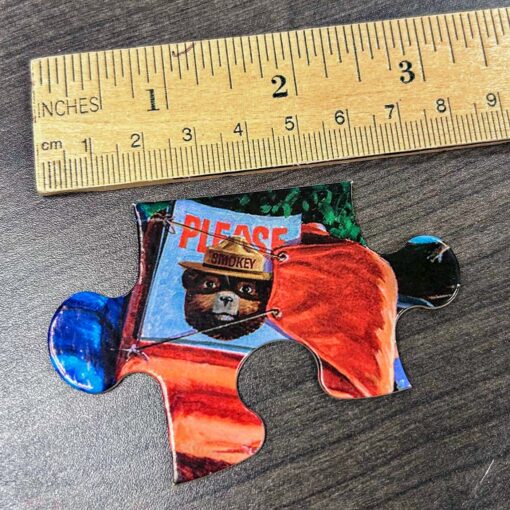 A single puzzle piece from the Smokey Bear Children's Puzzle placed next to a wooden ruler for size comparison. The piece features part of Smokey Bear holding a red sign and measures approximately 2 inches in width, designed for small hands to easily grasp.