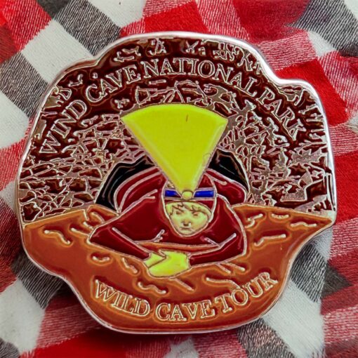 Wind Cave National Park Wild Cave Tour enamel pin displayed on a red and black flannel shirt. The pin depicts a caver with a glowing yellow headlamp navigating through tight cave formations, emphasizing adventure and exploration.