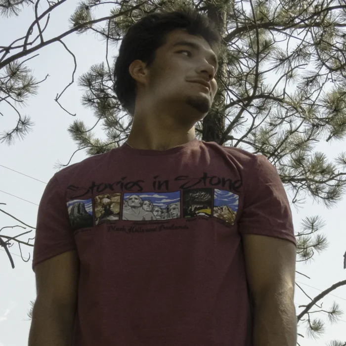 Man wearing red Stories in Stone shirt near a pine tree