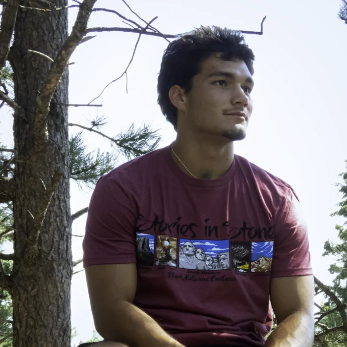 Man wearing red Stories in Stone shirt while sitting near a pine tree