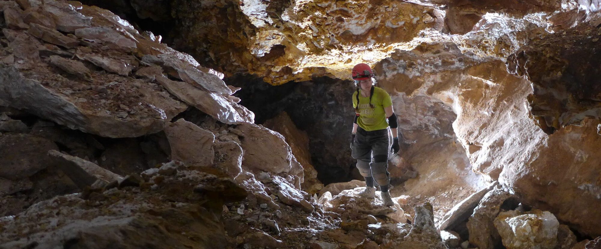 A caver in a yellow shirt explores a rocky cave passage