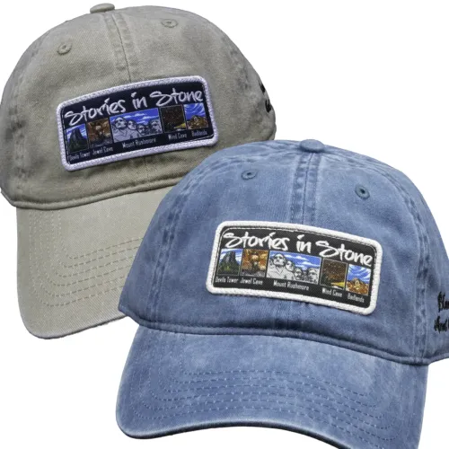 Two Stories in Stone baseball caps, one khaki and one denim blue, each with an embroidered patch showing colorful scenes of Devils Tower, Jewel Cave, Mount Rushmore, Wind Cave, and Badlands, with "Stories in Stone" stitched across the top.