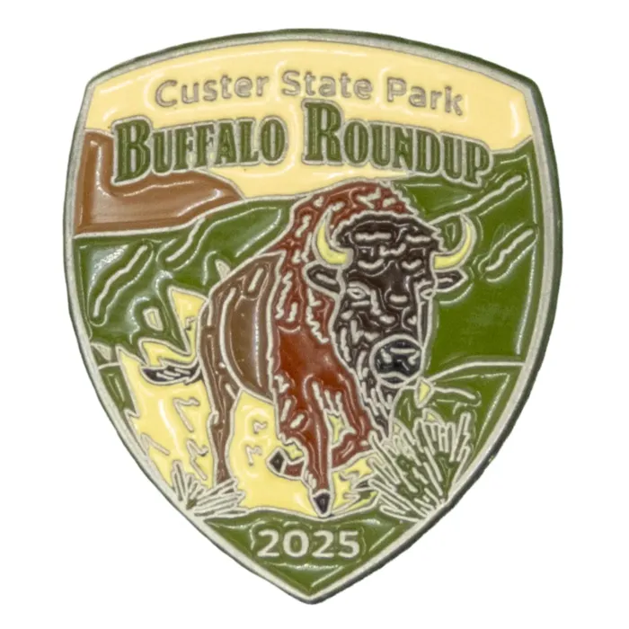 Close-up of the 2025 Custer State Park Buffalo Roundup enamel pin, showing a brown buffalo charging across yellow grass with green hills in the background. The text reads "Custer State Park Buffalo Roundup 2025."