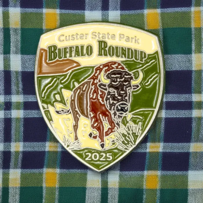 The 2025 Custer State Park Buffalo Roundup enamel pin photographed on a green, navy, and gold plaid shirt background. The pin features a buffalo design, green hills, and "2025" at the bottom.
