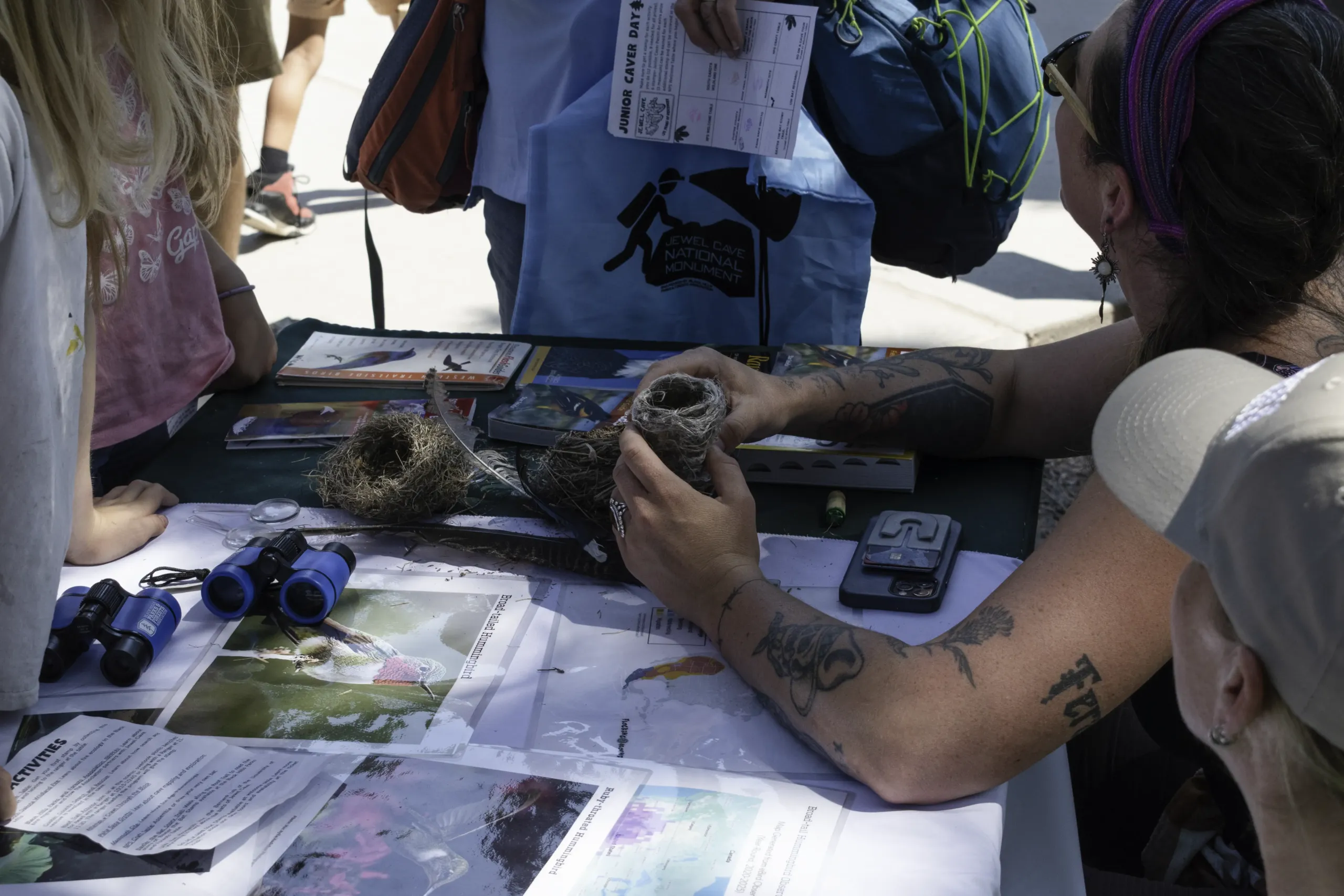 Children and volunteers examine bird nests, feathers, and field guides at a Junior Caver Day activity table.