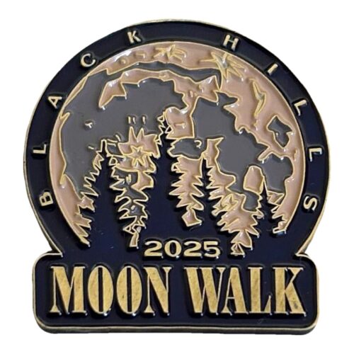Arched enamel lapel pin in navy blue and antique gold. A large full moon fills the arch behind a row of pine trees in silhouette. The letters BLACK HILLS curve around the top edge, and a banner across the bottom reads MOON WALK with the date 2025 above it.