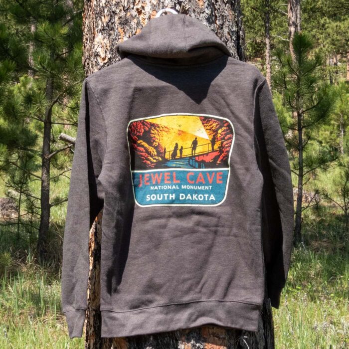 Charcoal Jewel Cave National Monument full-zip hoodie outdoors in the Black Hills, showing the back cave-tour sign